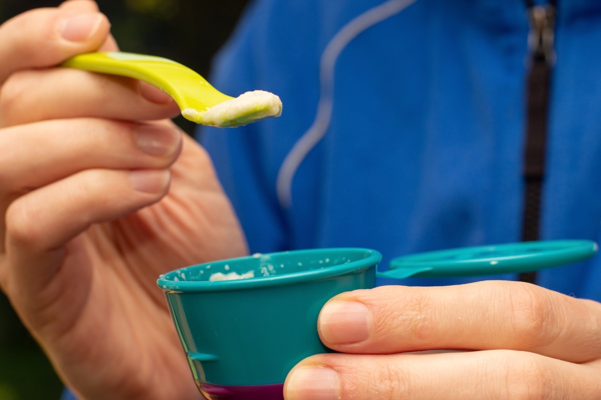 Fathers hand feeding little baby first solid food. Holding a spoon with rice cereal porridge and baby food container