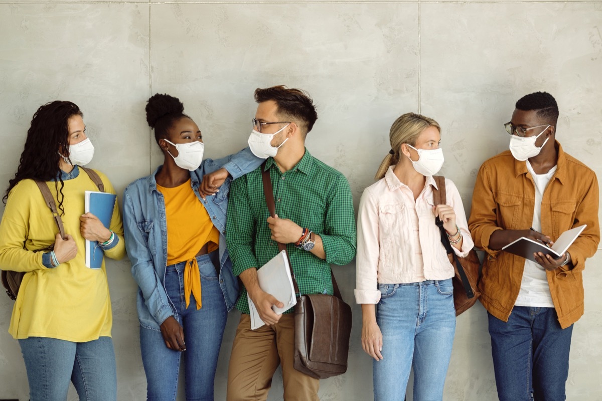 group of college friends communicating while wearing protective face masks while standing against the wall in a lobby.