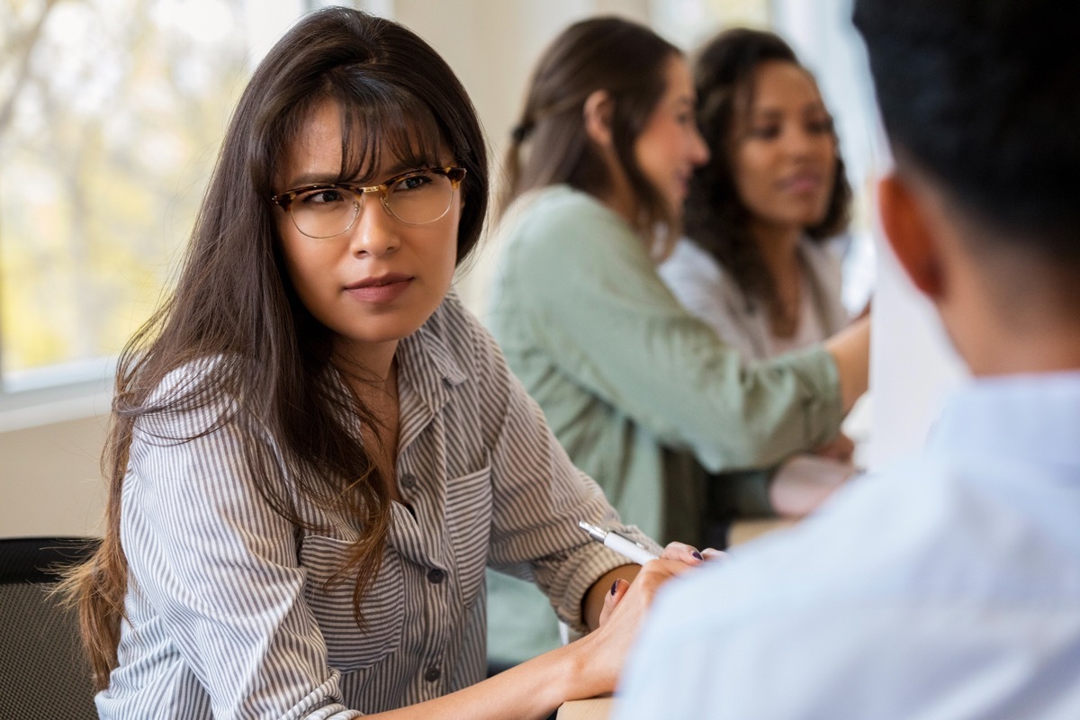 A serious young female creative professional sits at a conference table during a staff meeting next to an unrecognizable coworker. She frowns in disbelief as she listens to him.