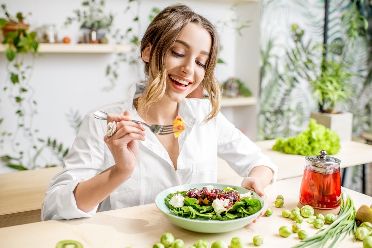 Young woman eating healthy food sitting in the beautiful interior with green flowers on the background