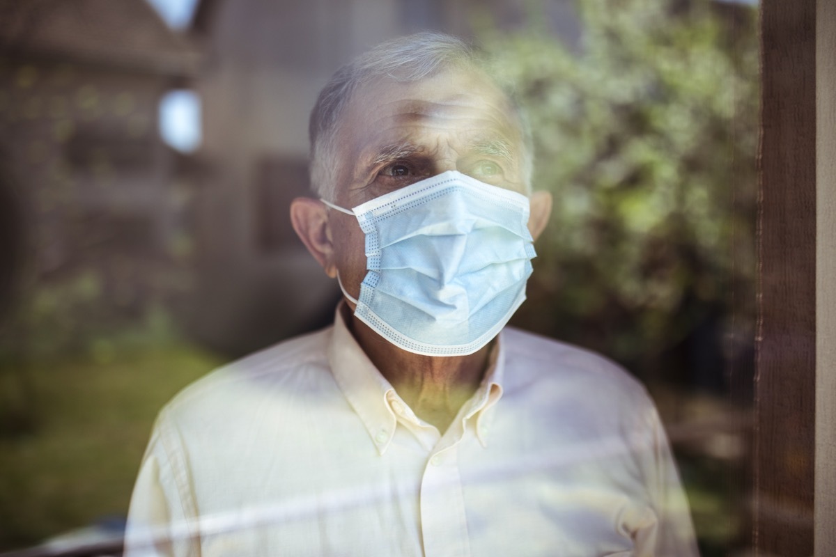 Senior man stays home because of Coronavirus. He stands by the window and looks out at the garden.