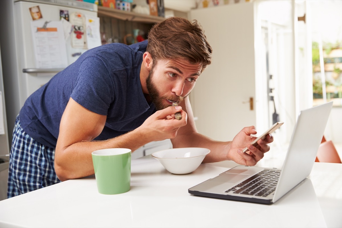 Man multitasking in the kitchen eating, standing, watching laptop, holding phone