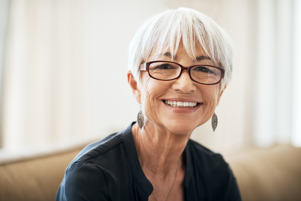 Cropped portrait of a senior woman sitting on the sofa at home. She has short white hair and is wearing glasses while smiling.