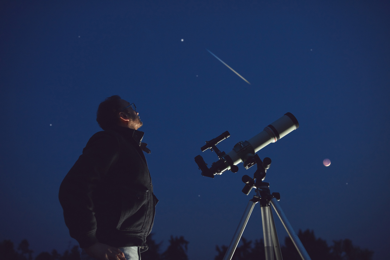 The silhouette of a person looking up at the night sky with a telescope