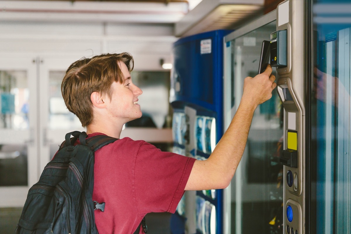 Student teenage boy wearing backpack uses mobile phone to pay for snack and drink at vending machine