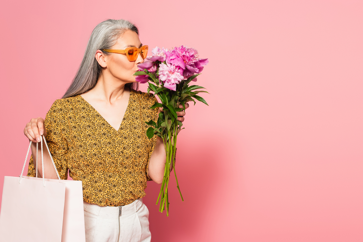 mature woman with shopping bag enjoying aroma of fresh peonies on pink background