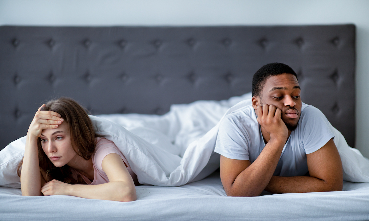 A young couple under the covers at the foot of the bed, each looking away from each other and upset.