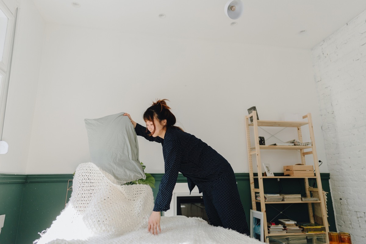 small bedroom ideas - Photo of a young woman making her bed right after waking up, still in her PJ's, arranging bedsheets and pillows.