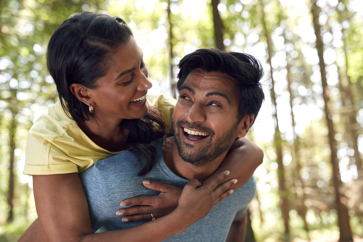 Portrait Of Couple Hiking Or Walking Through Woodland With Man Giving Woman Piggyback