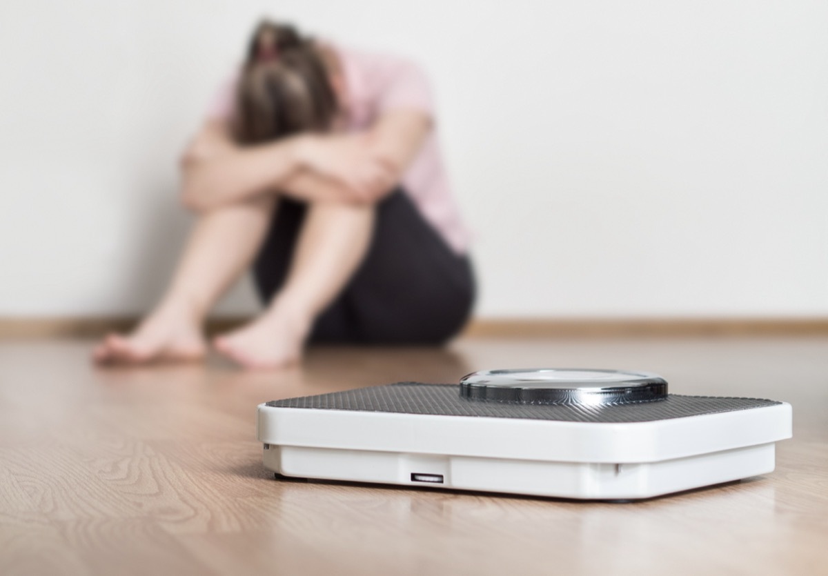 woman sitting on floor holding head and arms on knees with a scale in the foreground