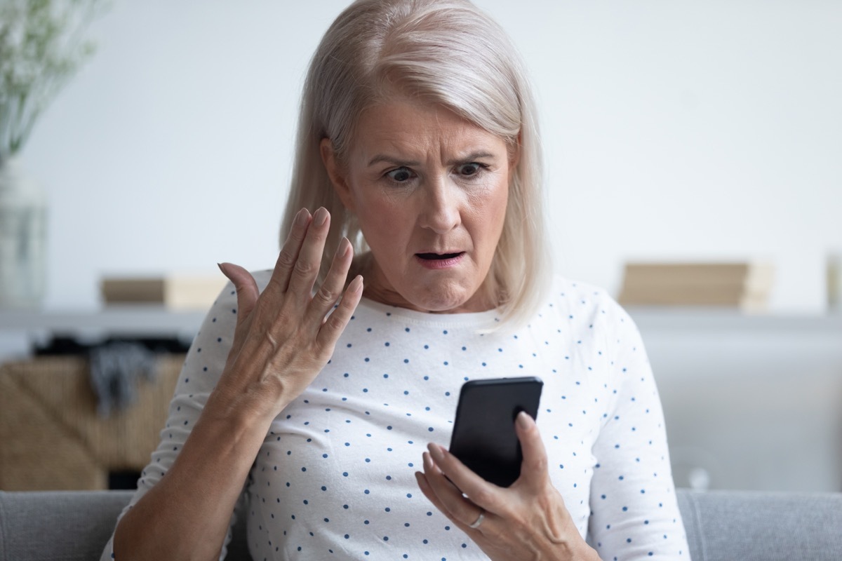 Elderly 50s woman sitting on couch in living room holding smart phone gesturing looking annoyed feels angry having problem with gadget, slow internet, connection lost, discharged broken device concept