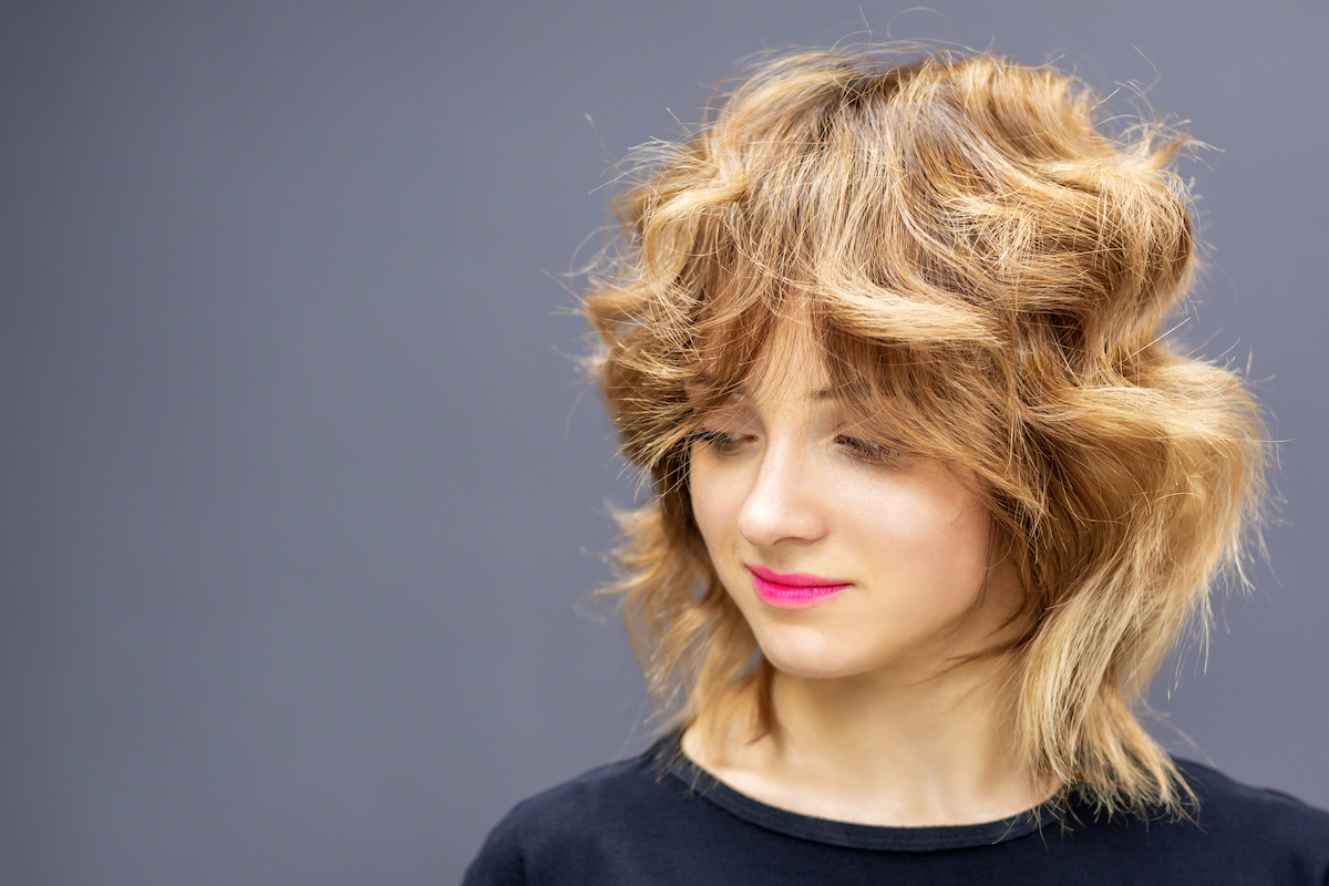 Blonde woman with thick wolf-cut hairstyle wearing a black top against a gray background