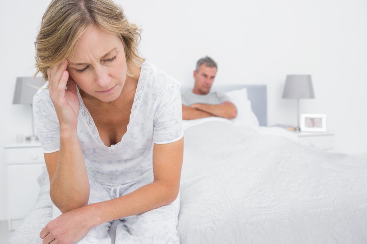 older woman looking upset in bed while man sits behind her