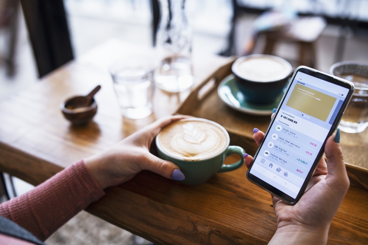 Young woman checking banking account via mobile app on smartphone while drinking coffee at Cafe.