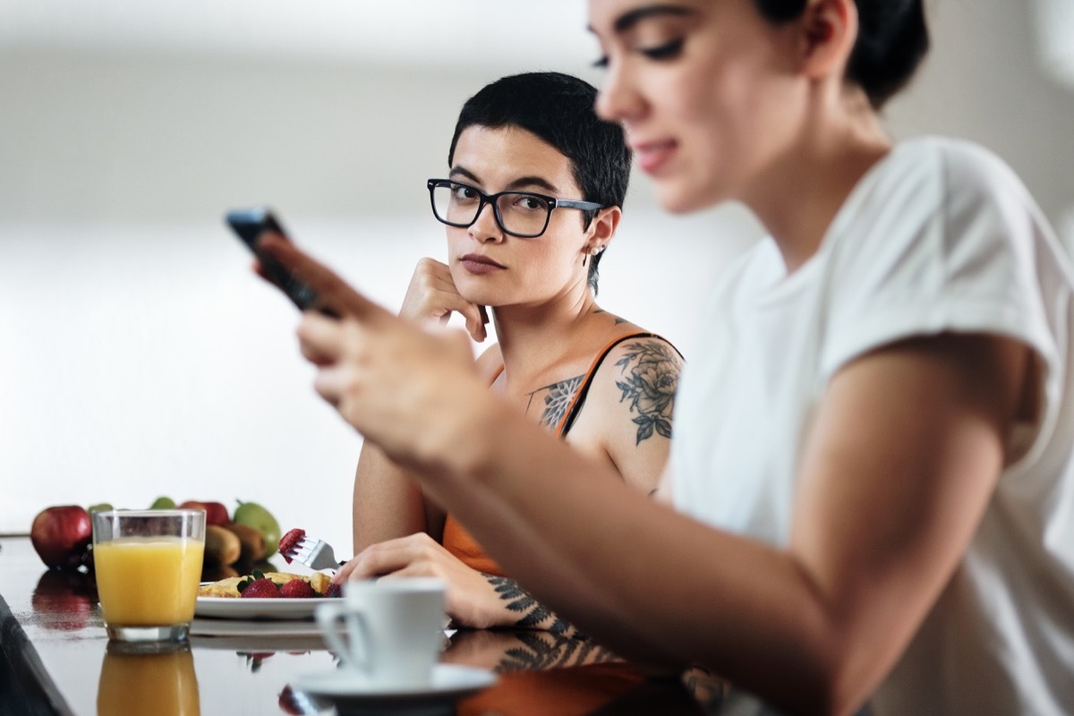 two lesbian women at home eating breakfast, partner chatting on mobile telephone. Young woman being ignored by her girlfriend and feeling jealous