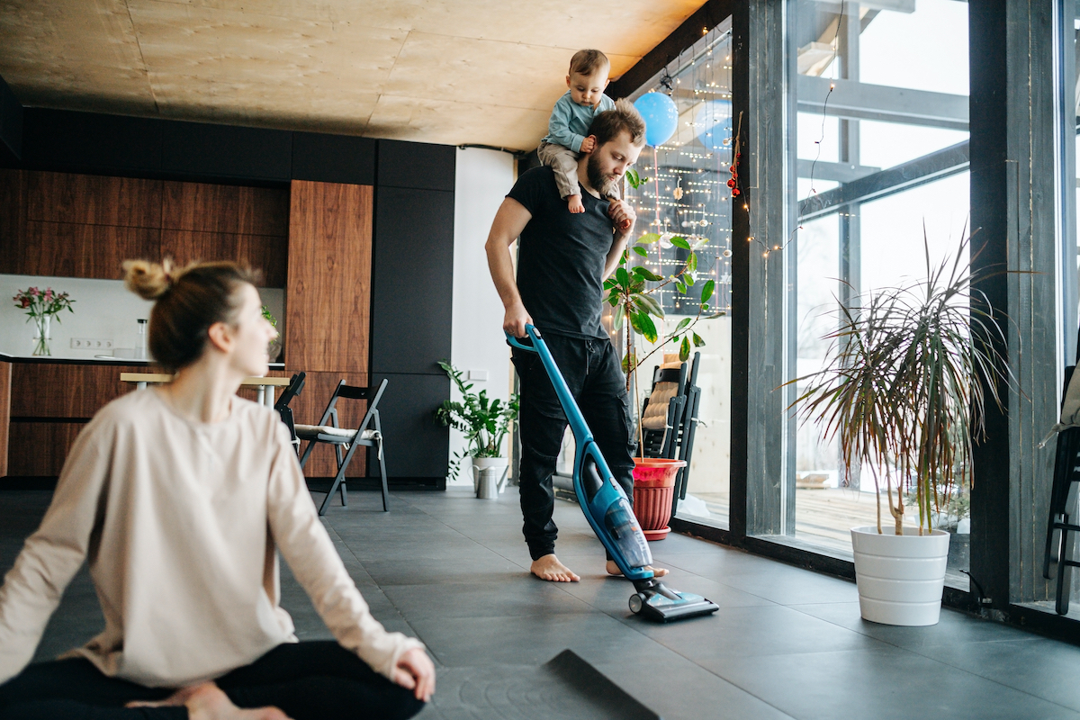 Mother sits at easy simplified lotus pose, looking at the father vacuum cleaning apartment floor with their infant baby riding on his neck