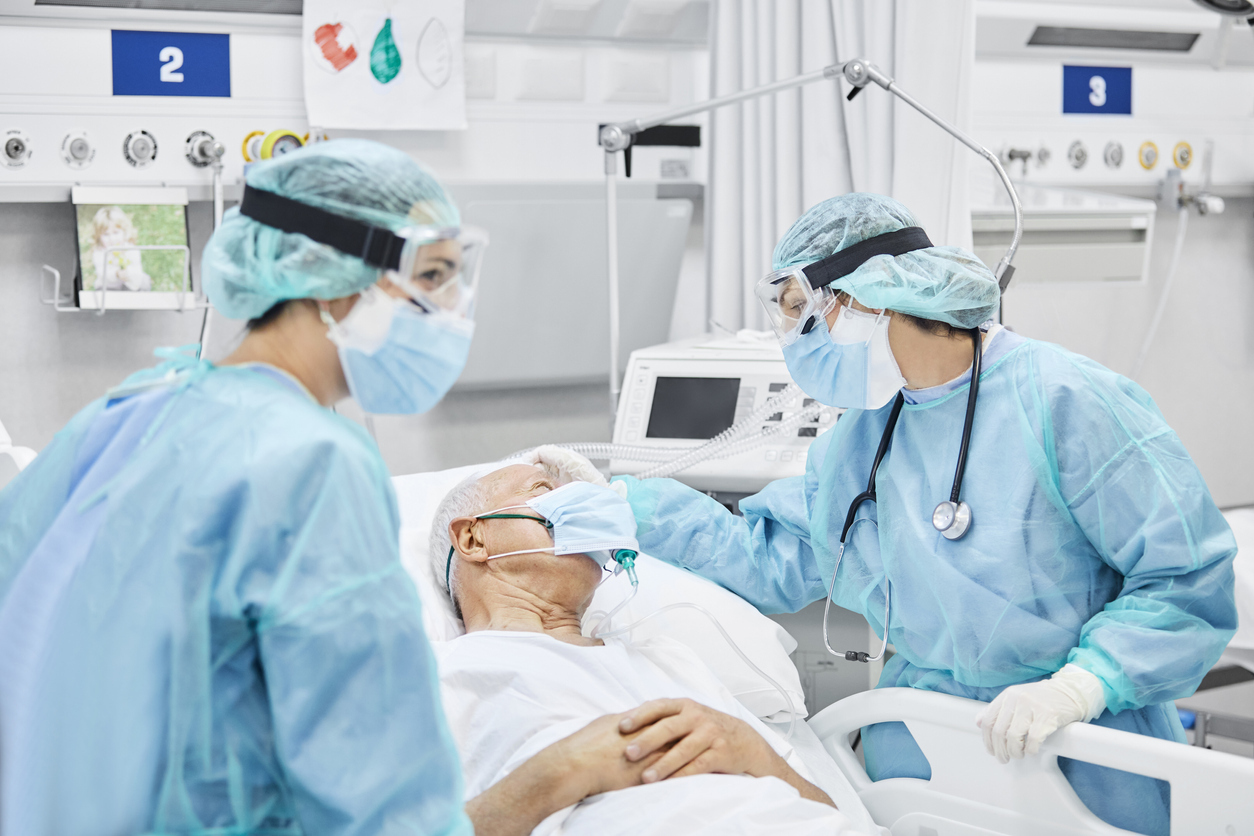 Two female doctors wearing full protective gear check on an older male patient in the ICU for coronavirus