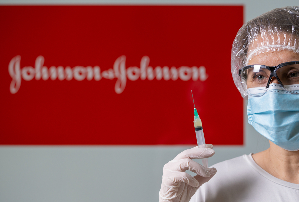 A woman wearing protective glasses, a face mask, and a hair covering holds a syringe in front of a Johnson & Johnson sign