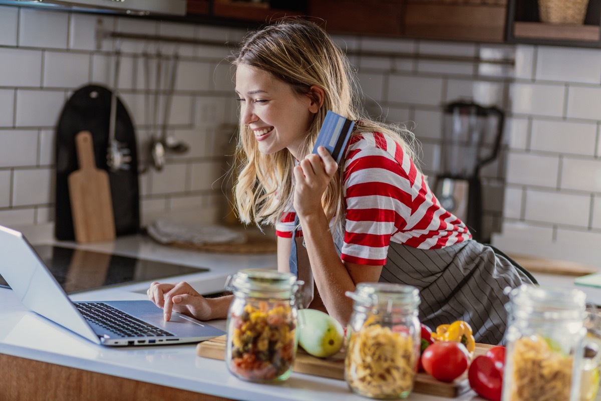 A young beautiful woman is in the kitchen, she prepares a meal while using a laptop and credit card for online buying