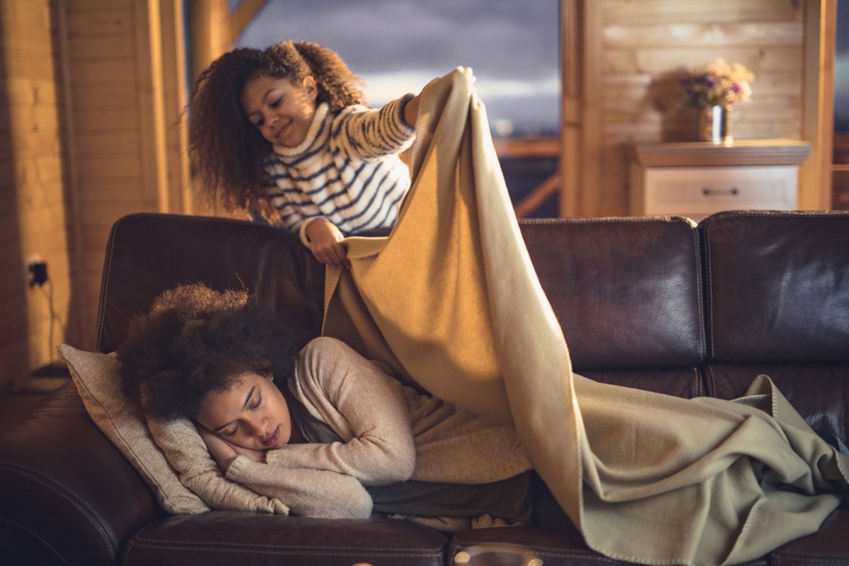 girl covering mother with blanket while she is sleeping on sofa at home. Focus is on woman.