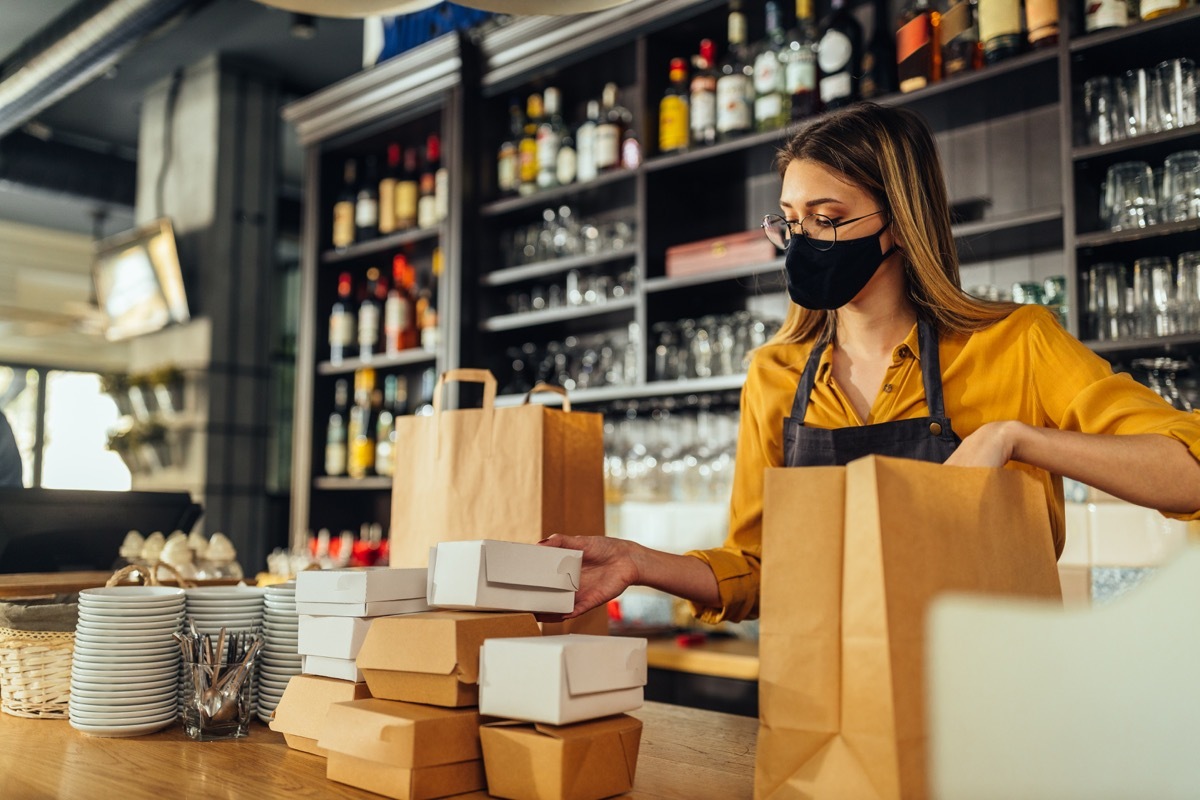 Fast food restaurant during coronavirus pandemic. Female fast food worker with protective face mask packing meals