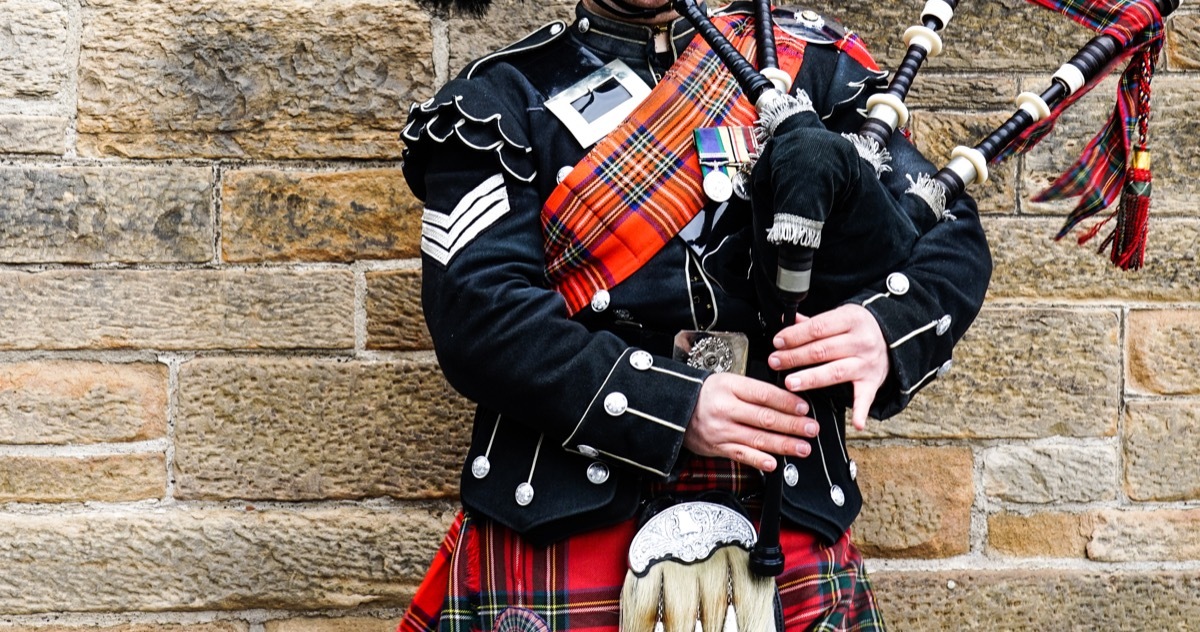 Scottish bagpiper dressed in traditional red and black tartan dress stand before stone wall, Prince Philip controversial moments