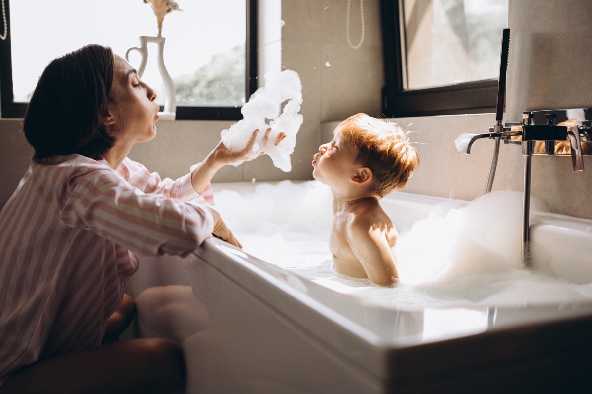 mother washing and blowing bubble with young son in bathtub