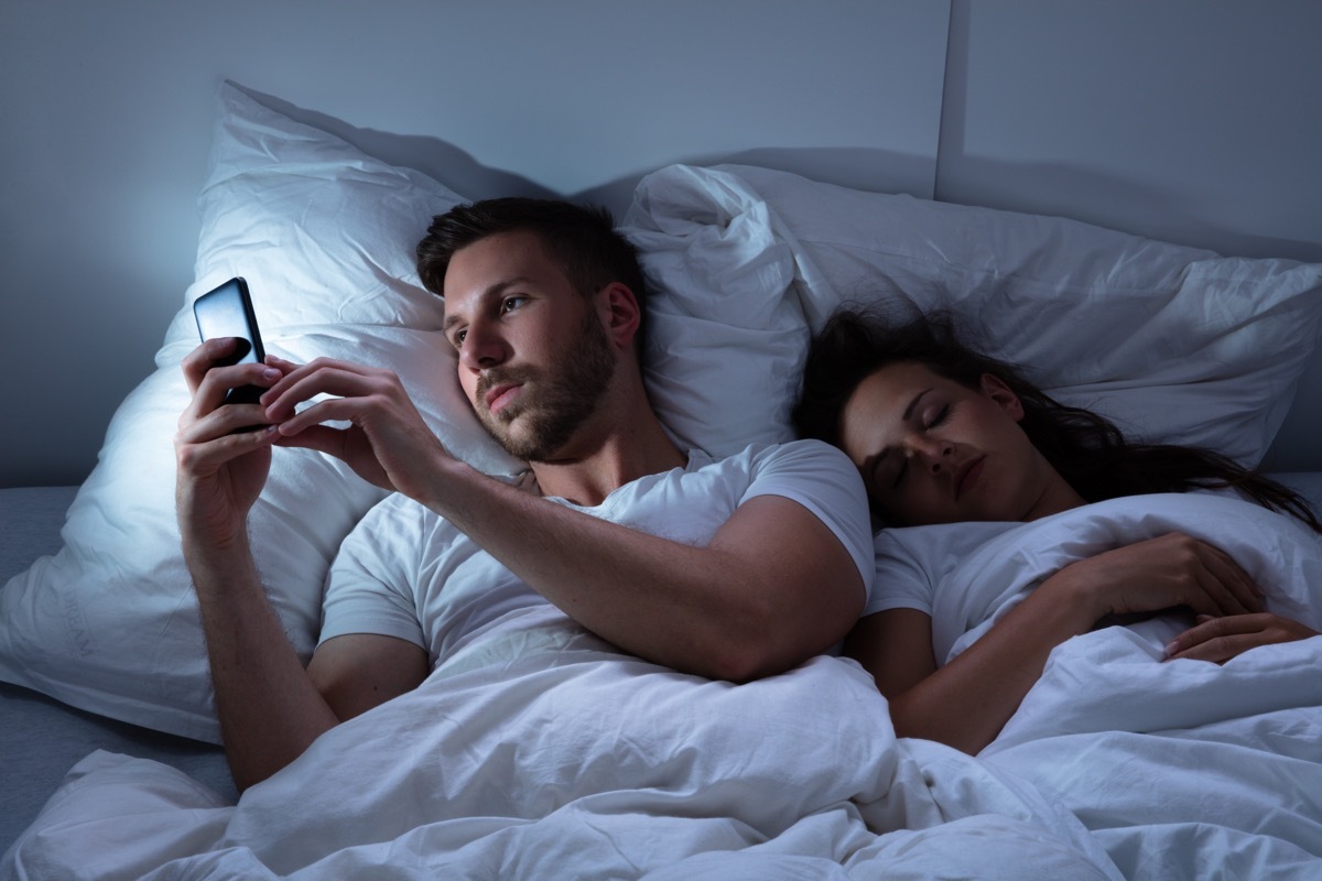 Man using his phone in bed while woman sleeps next to him, signaling cheating