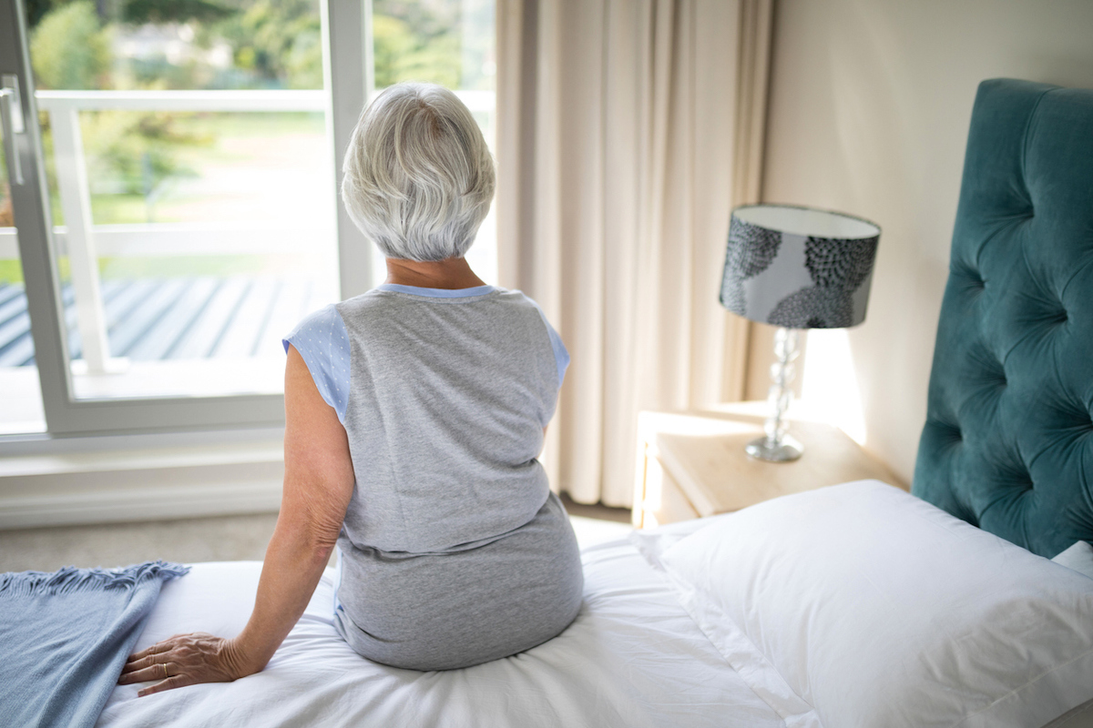 Rear view of senior woman sitting on bed in bedroom, looking out window