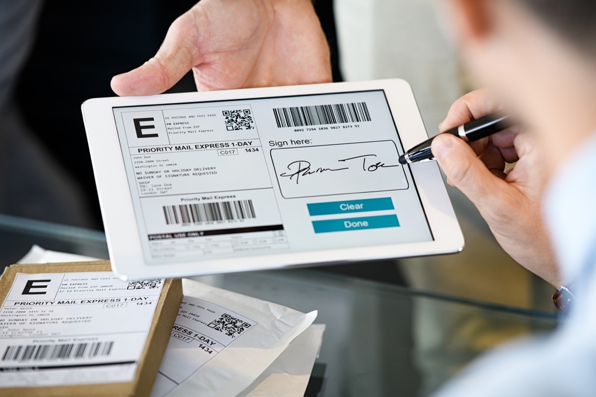 Close up hand of businessman signing on digital tablet while receiving parcel. Man signing delivery service document on tablet after receiving package. Signing digital document after receiving parcel from courier in office.