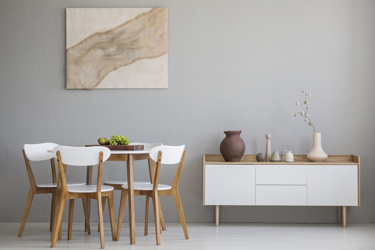 Real photo of wooden table and four chairs standing in light grey dining room interior with modern art poster and cupboard with decor