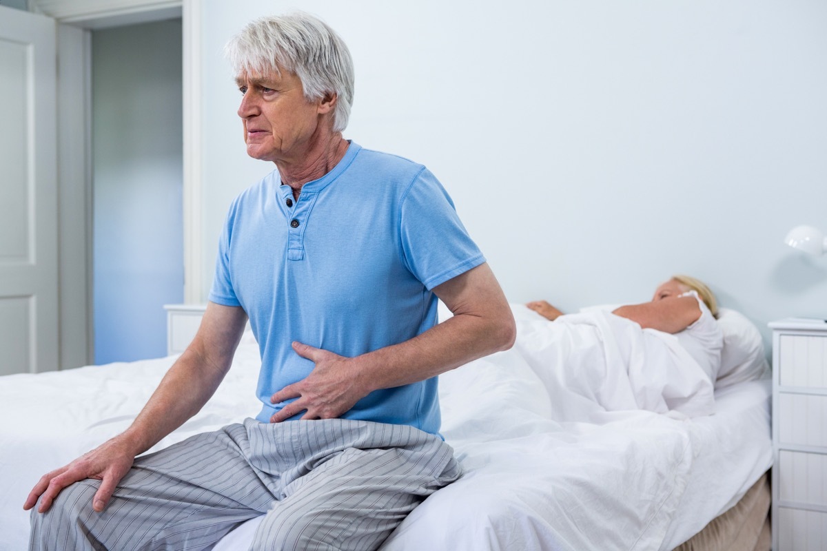 senior man with white hair sits at edge of bed holding stomach while wife sleeps, what your stomach pain could mean