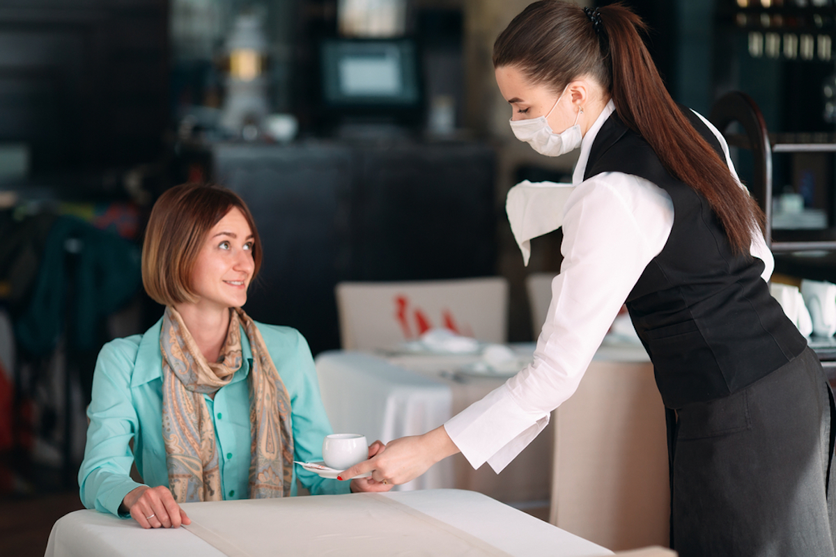 young woman with bob sitting at table in restaurant being served coffee by woman in mask