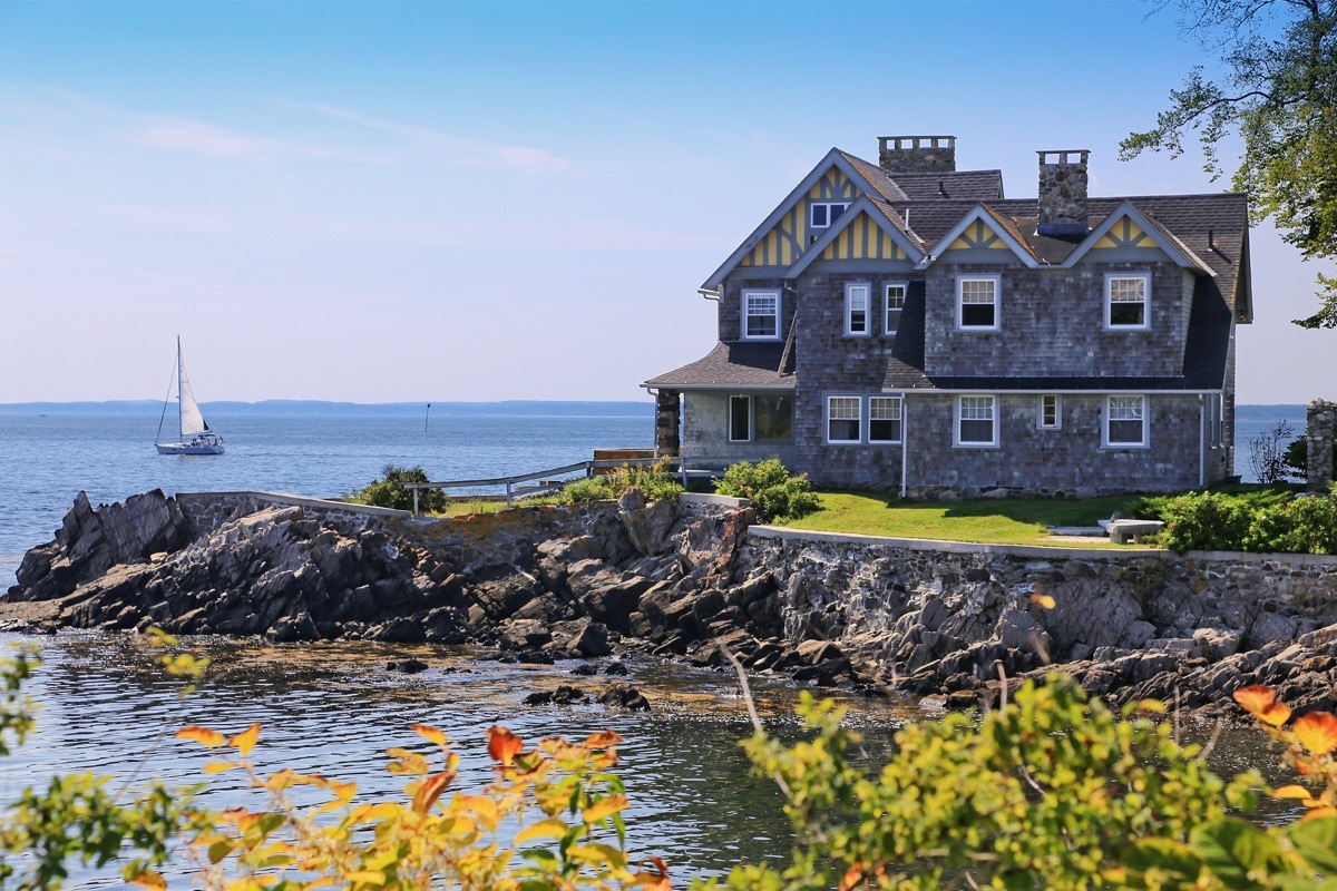 Luxury Waterfront House with grey shingle exterior, Kennebunkport, Maine, New England, USA. Rocky shore, ocean waters, sailboat, green and yellow bushes, trees, and blue sky with clouds are in the image.