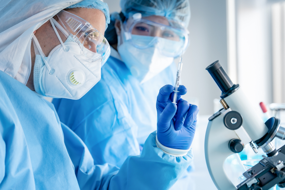 A pair of scientists working in a lab while wearing full protective gear, with one holding a syringe