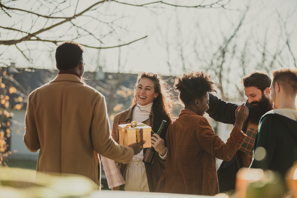 Candid shot of cheerful young couple welcoming and greeting their friends who came to their home as guests. They are standing outside, in warm autumnal sunshine.