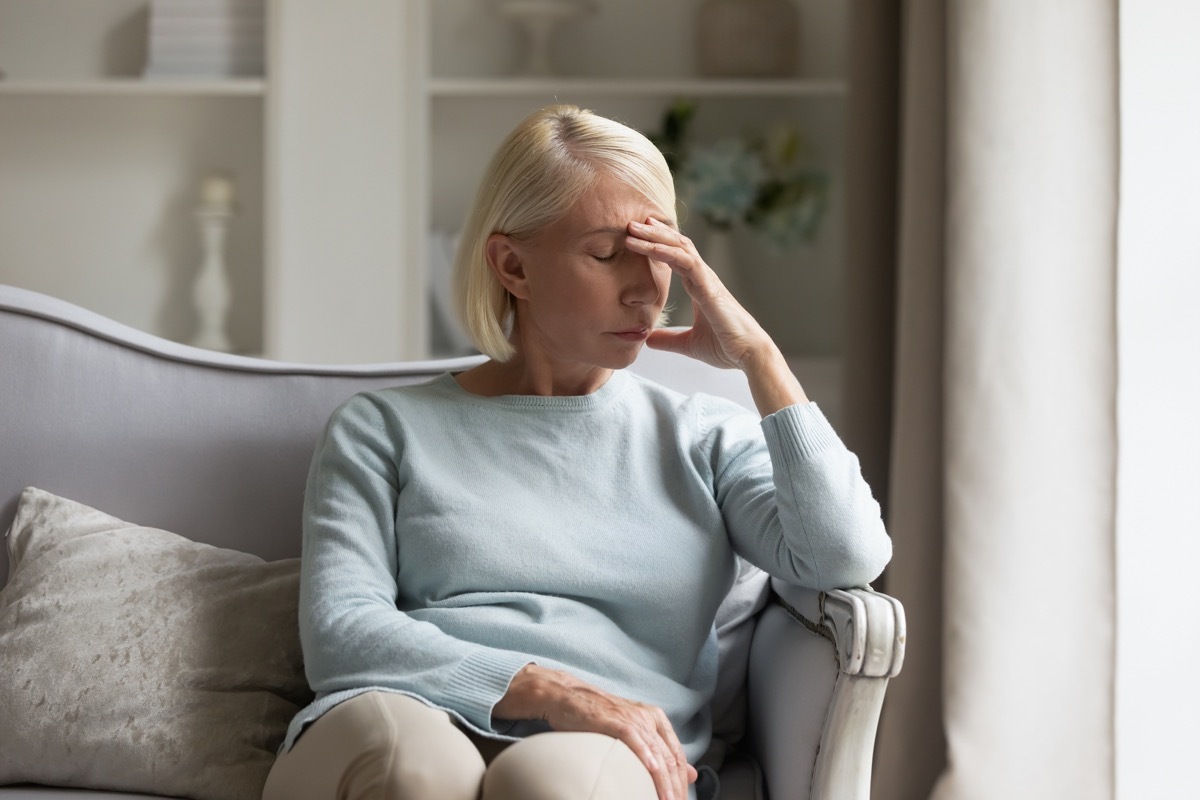 Tired older woman sitting on comfortable sofa in living room, touching forehead. Exhausted mature lady suffering from head ache at home. Unhappy elder grandmother worrying about bad news alone.