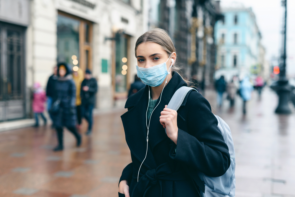 A teenage girl wearing a dark jacket, backpack, and face mask walks down a rainy city street.