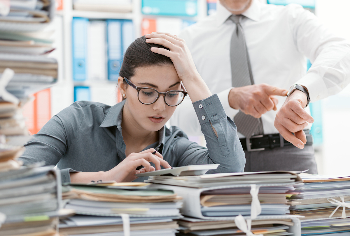 Demanding boss pointing to his watch and asking his employee to hurry up while she sits behind a stack of folders and papers