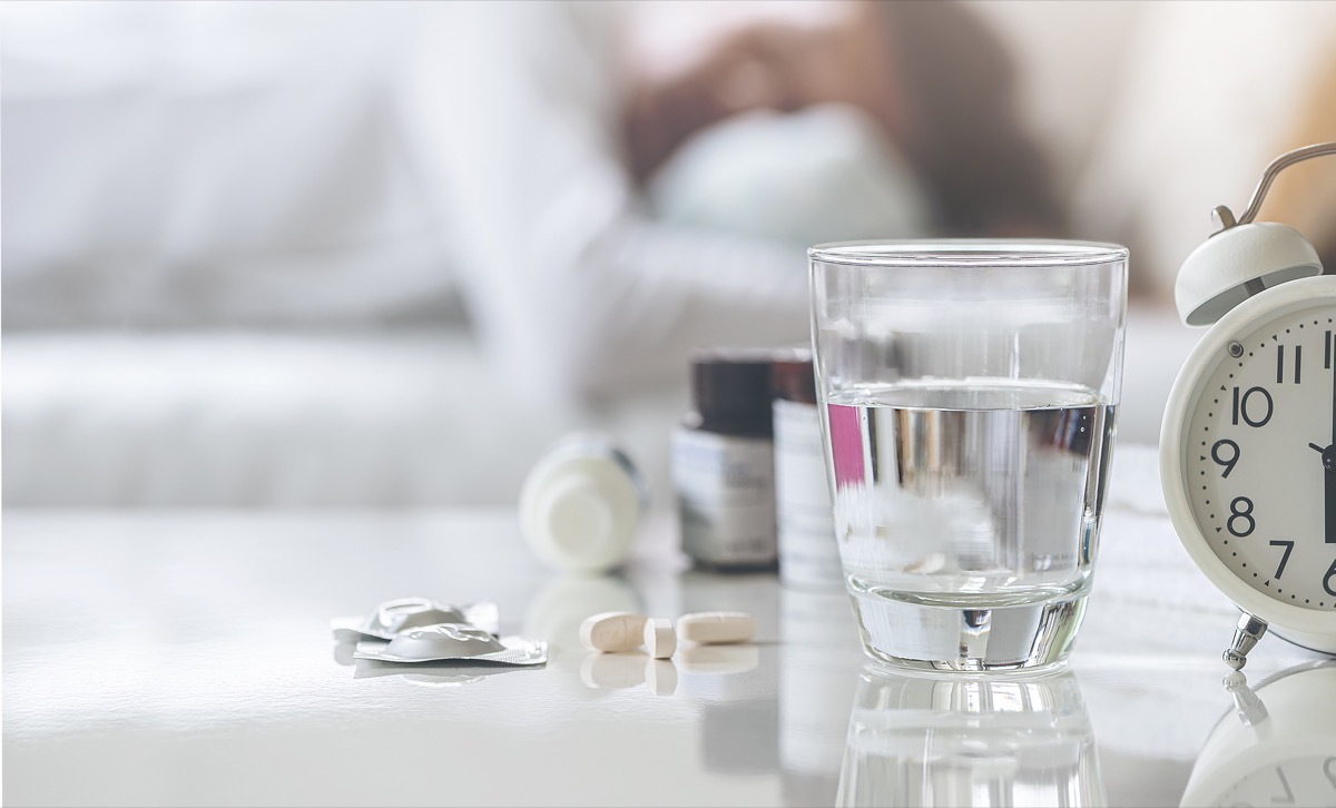 Closeup glass of drink water and pills on white table with blurred background of man sleeping on sofa, medicine and health care concept, copy space.Closeup glass of drink water and pills on white table with blurred background of man sleeping on sofa, medicine and health care concept, copy space.