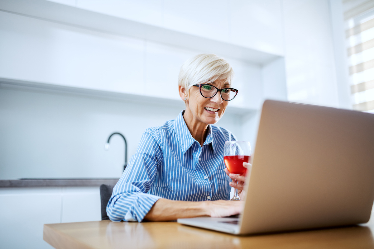 Happy smiling white blonde senior woman sitting at dining table, drinking red wine and having video call over at laptop