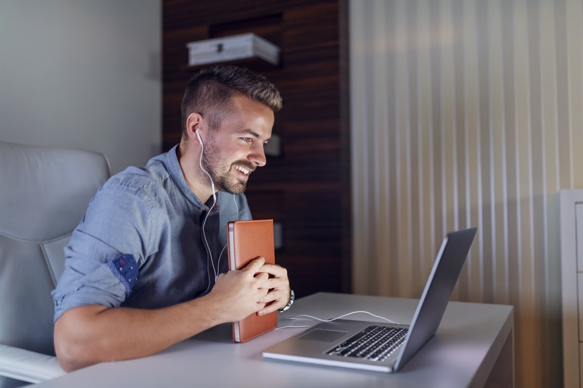 Handsome bearded employee holding agenda in hands, having video call over laptop and sitting in office late at night.