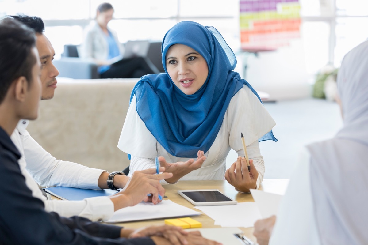 A serious Malaysian businesswoman sits at a table with unrecognizable clients and gestures as she speaks. There is a digital tablet on the table.