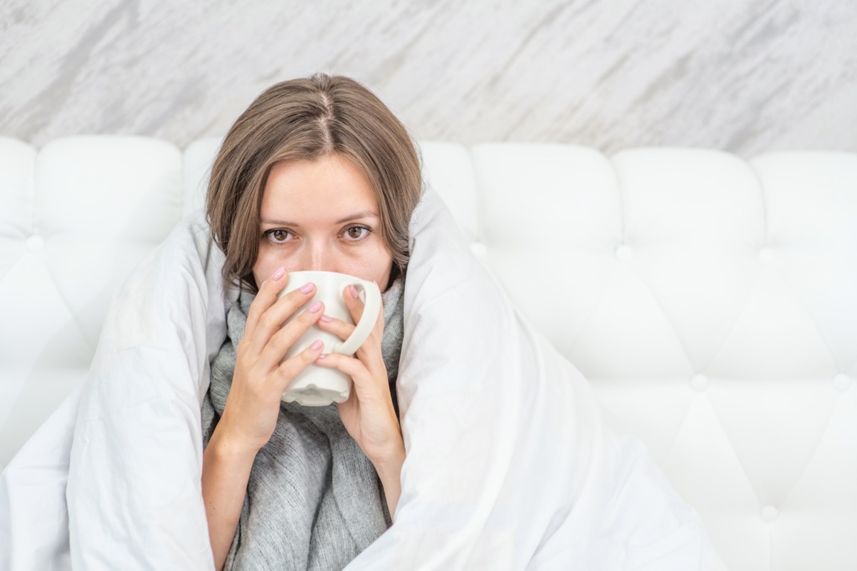 sick woman sitting on the bed wrapped in a white blanket and drink hot tea.