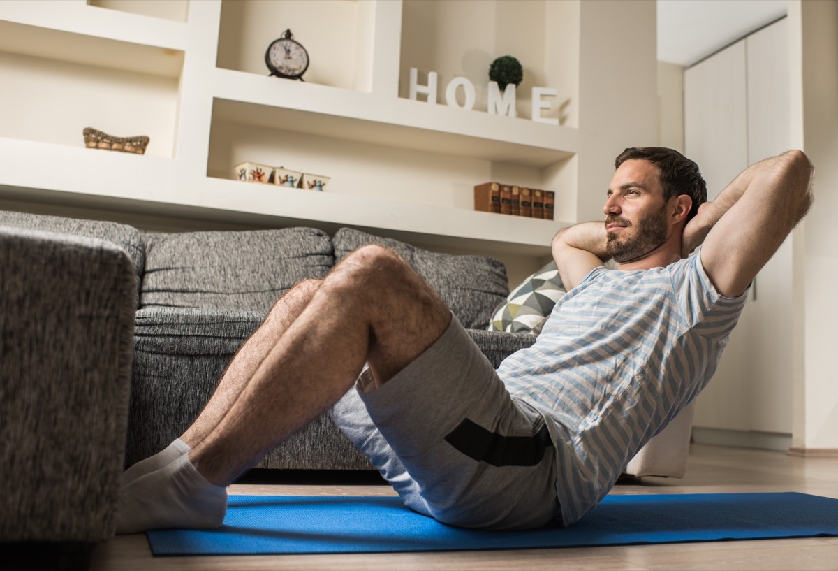 Athletic man sitting on exercise mat at home and exercising sit-ups with hands behind his head.