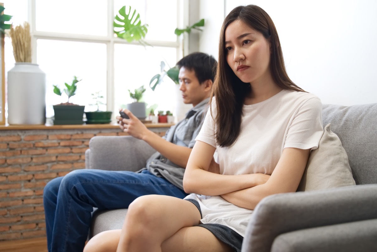 young couple sitting on a couch, apart from each other, girl looking upset and man on phone 