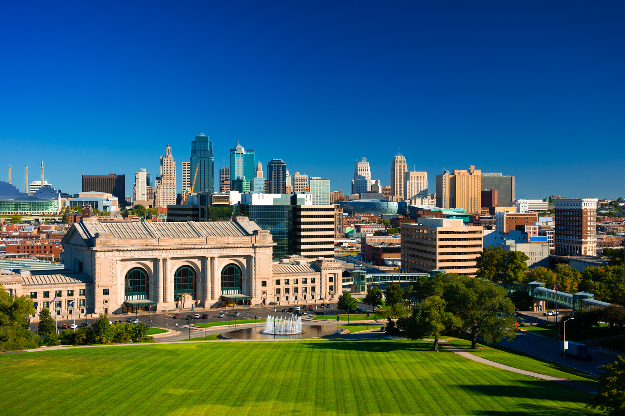 The skyline of Kansas City Missouri with a deep blue sky in the background and Union Station, a fountain, and Penn Valley Park in the foreground.