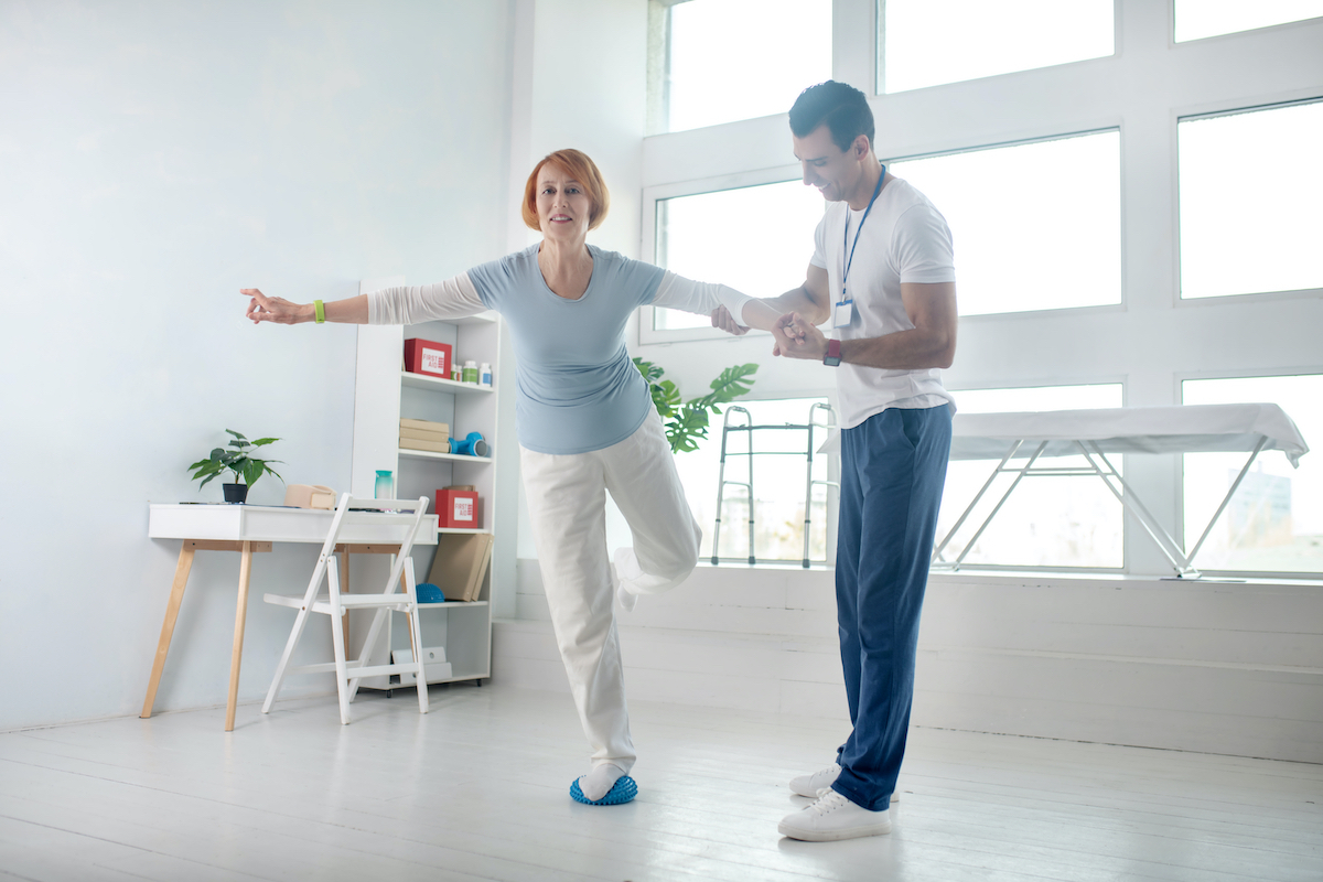 Balance exercise. Positive aged woman standing on one leg while holding the hand of her instructor