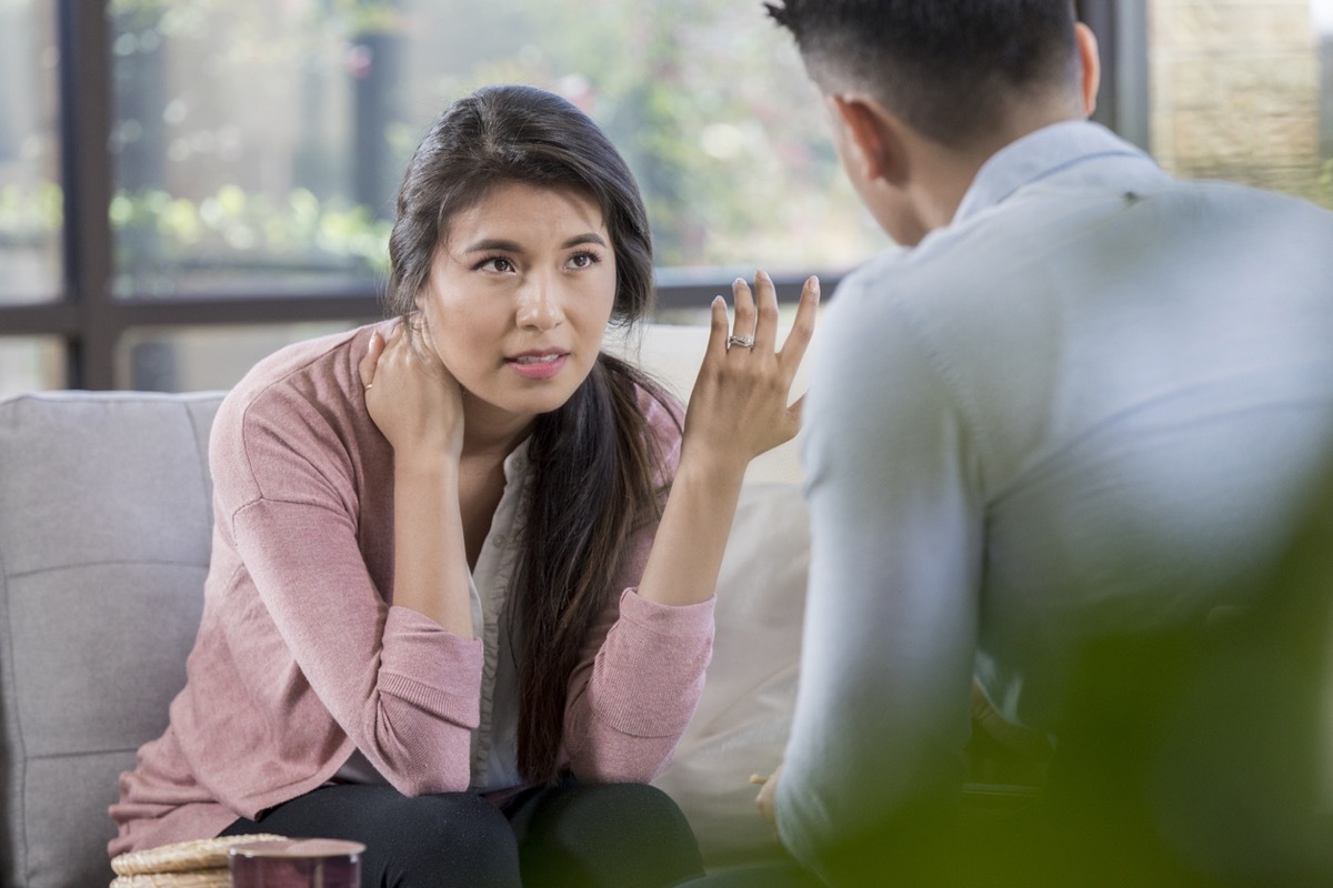 While in a disagreement, a young couple discusses problems. The wife sits on the sofa and gestures in frustration while husband listens intently